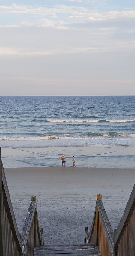 Wooden beach-access steps leading down to the sand, with two people at the water's edge.