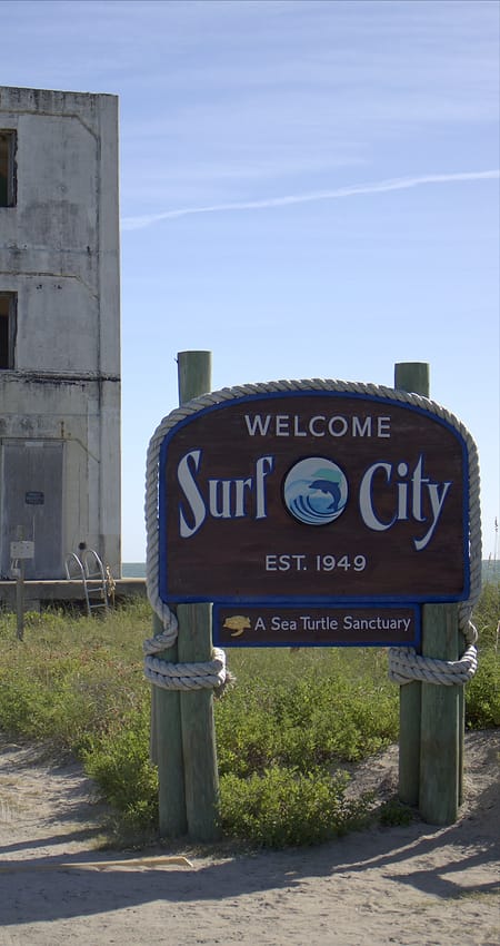 The Welcome to Surf City sign on a sandy path, with a weathered concrete tower behind it.