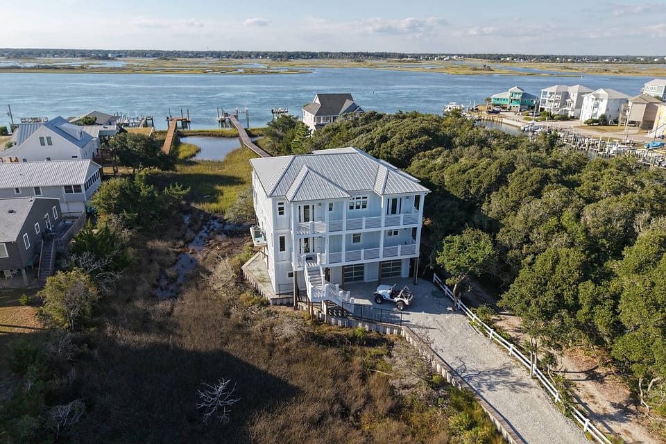 An aerial view of a white three-story coastal house on pilings, with docks reaching into the sound behind it.
