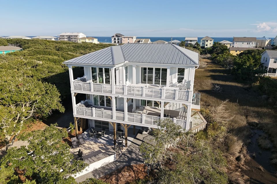 The back of a white coastal house from the air, two tiers of covered porches above a patio, the ocean beyond.